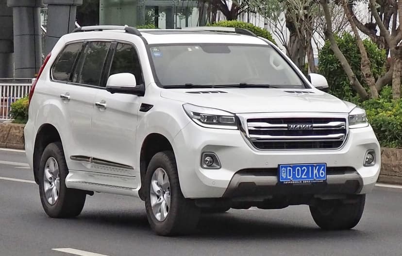White Haval H9 SUV driving on a road with green trees and urban buildings in the background.