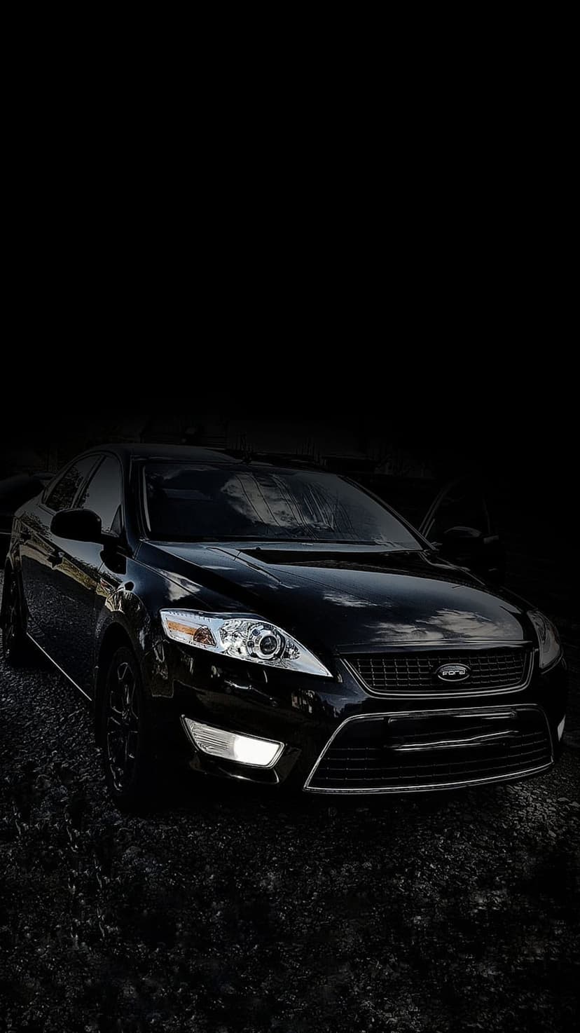 Black Ford Mondeo sedan front view with glowing LED headlights and a chrome grille against a dark night sky.