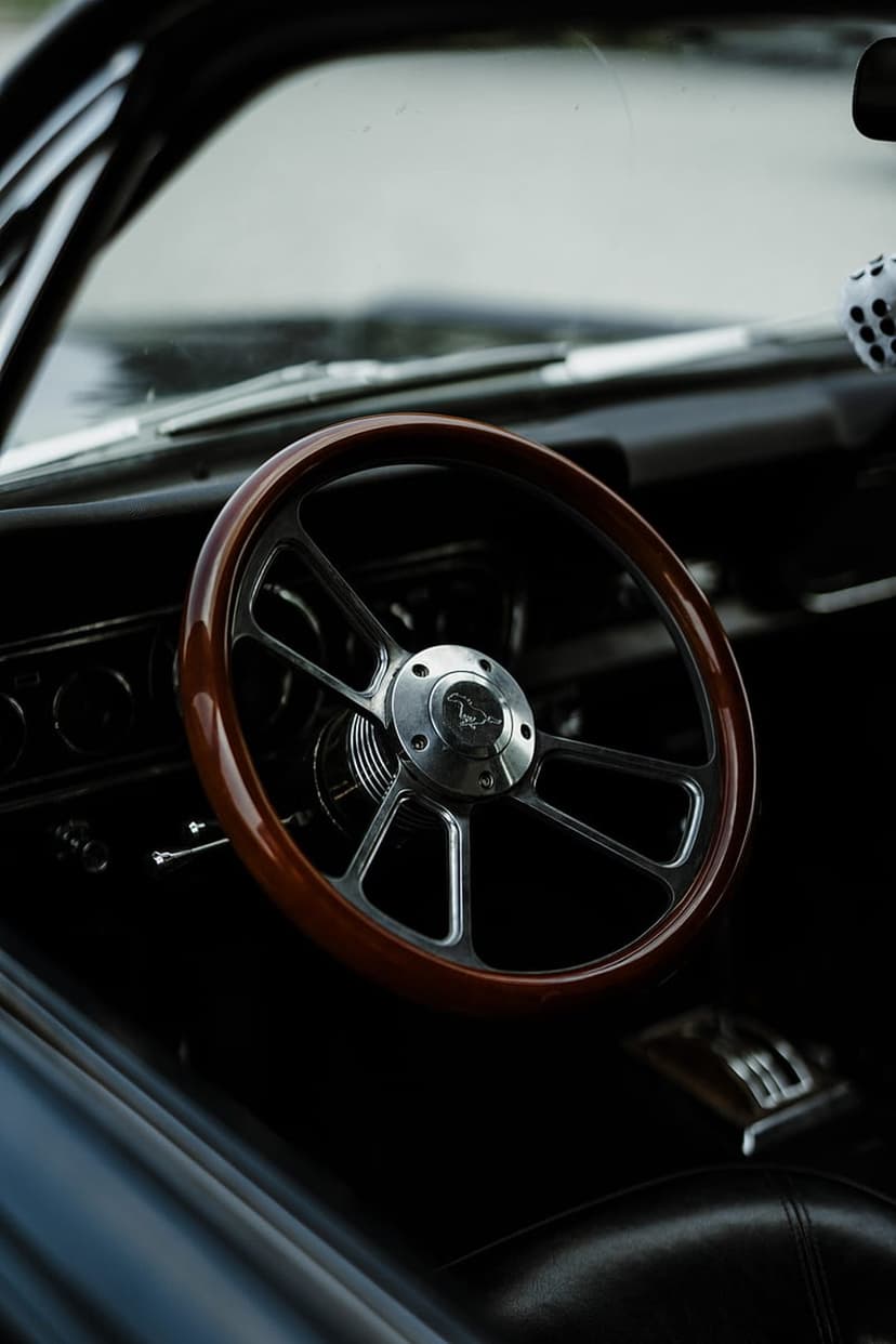 Polished wooden steering wheel with a chrome hub and white fuzzy dice hanging inside a vintage car interior.