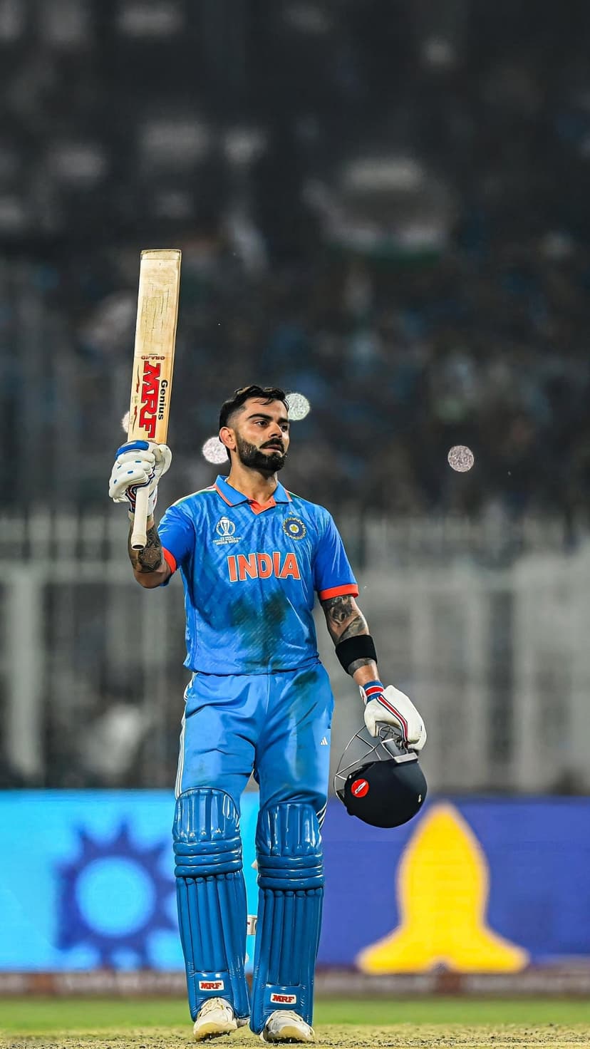 Virat Kohli wearing a blue Indian jersey holds a cricket bat and helmet against a blurred stadium background.