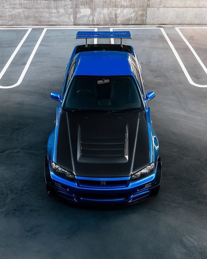 Blue Nissan Skyline R34 GT-R with a black carbon fiber hood parked on asphalt viewed from an aerial perspective.