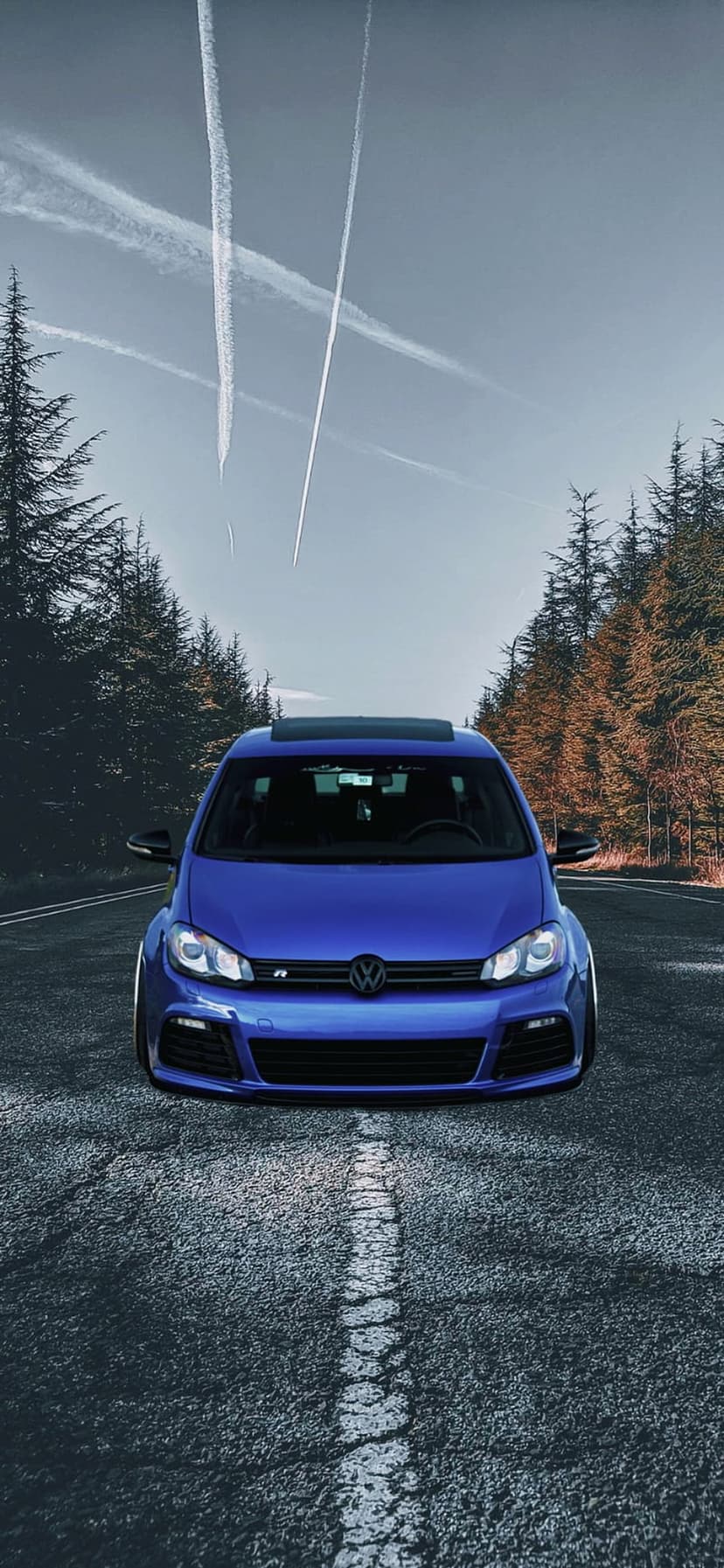 Blue Volkswagen Golf R parked on a cracked forest road with tall pine trees and jet trails in a pale blue sky.