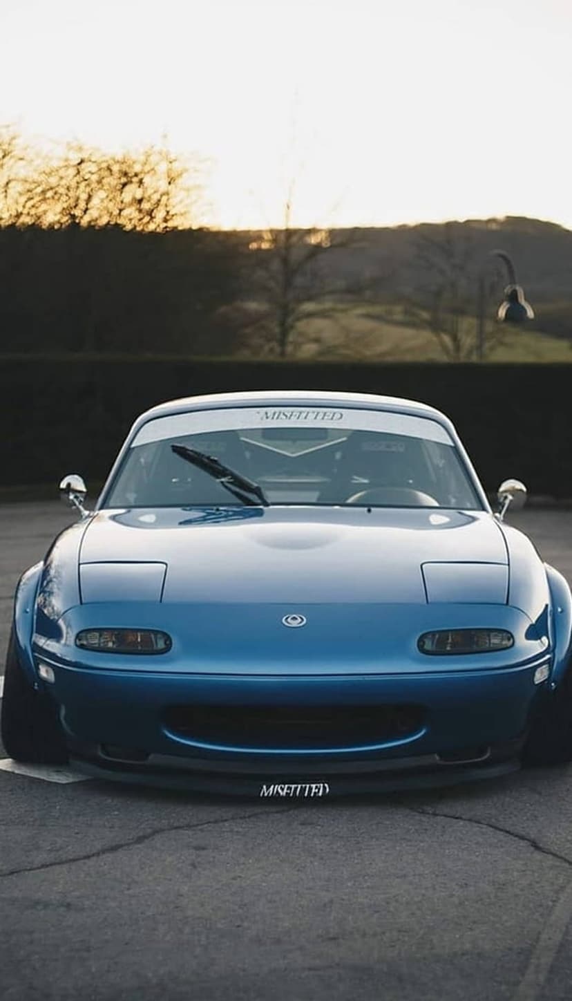 Blue Mazda Miata with wide fenders and pop-up headlights parked on asphalt during a golden hour sunset.