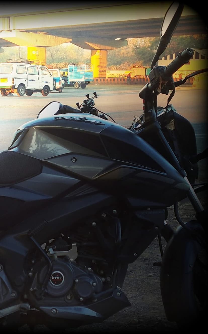 Dark motorcycle engine and fuel tank reflecting sunlight with blurred trucks under a concrete city overpass.