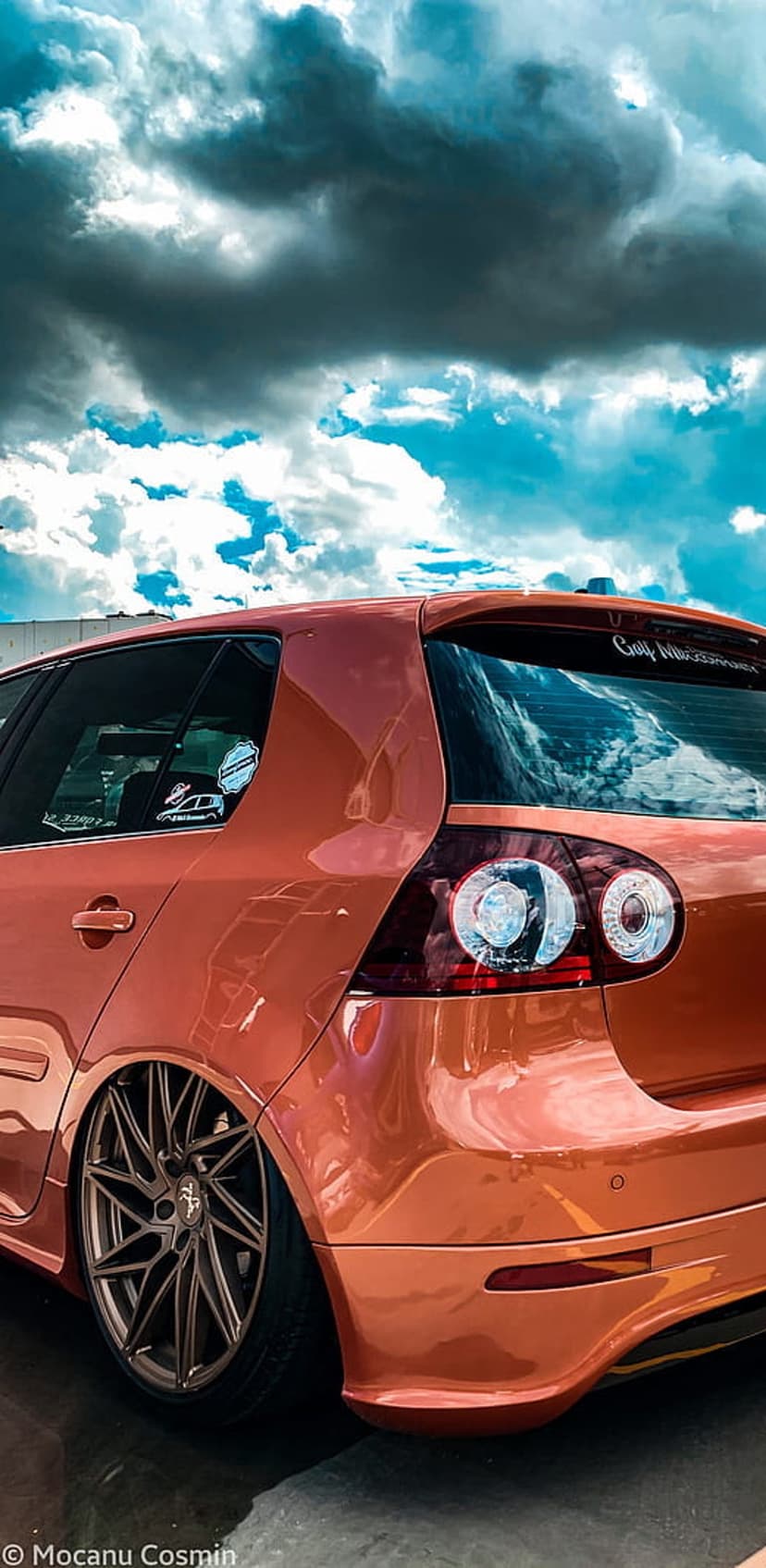 Orange Volkswagen Golf with extreme lowered stance and split-spoke rims under a dramatic overcast sky.