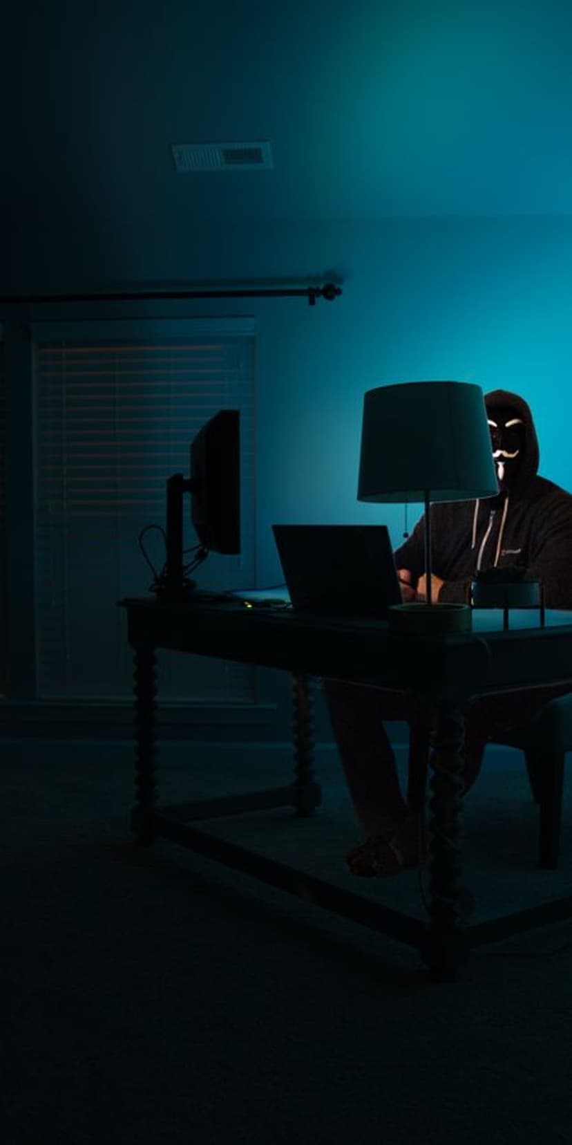 Guy Fawkes mask worn by a hooded person sitting at a laptop computer illuminated by a moody blue ambient glow.