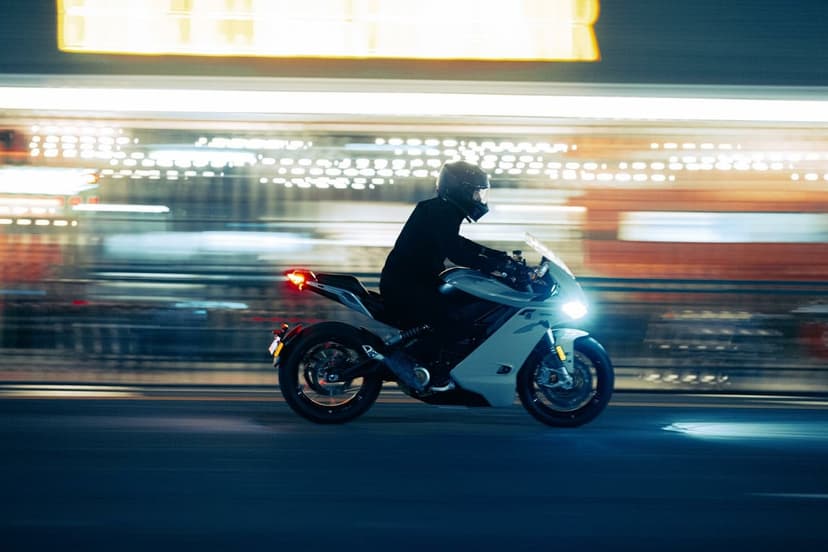 Motorcycle rider on a light grey sportbike speeding past blurred city lights at night with bright headlights.