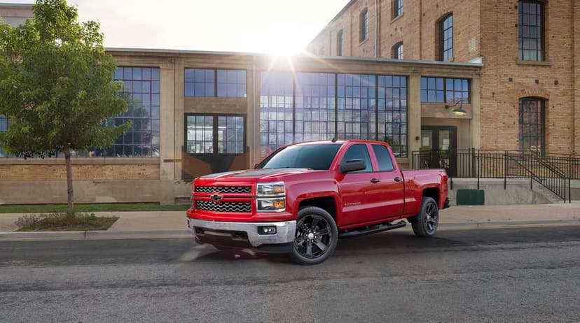 Red Chevrolet Silverado pickup truck with black rims parked in front of a brick factory building with sun flare.