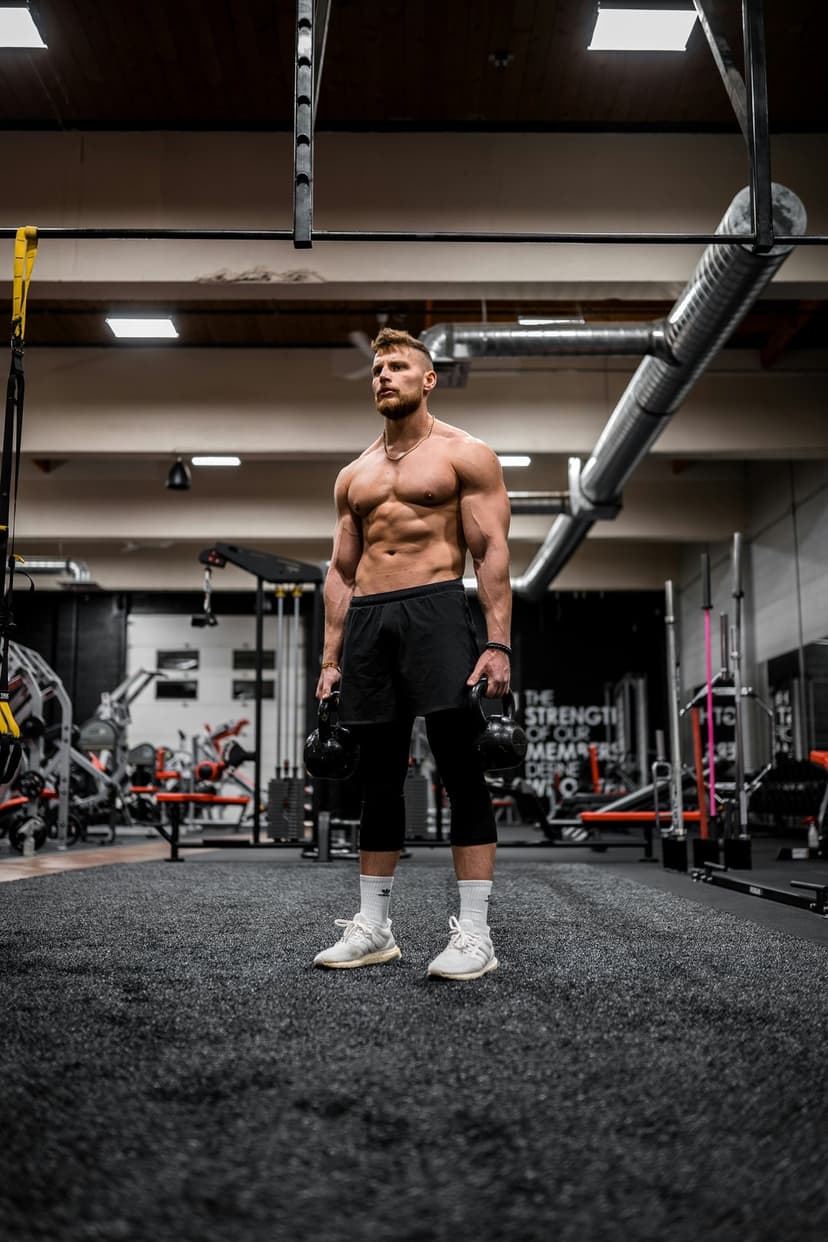 Muscular man with defined abs holding two kettlebells in a gym while wearing black shorts and white sneakers.