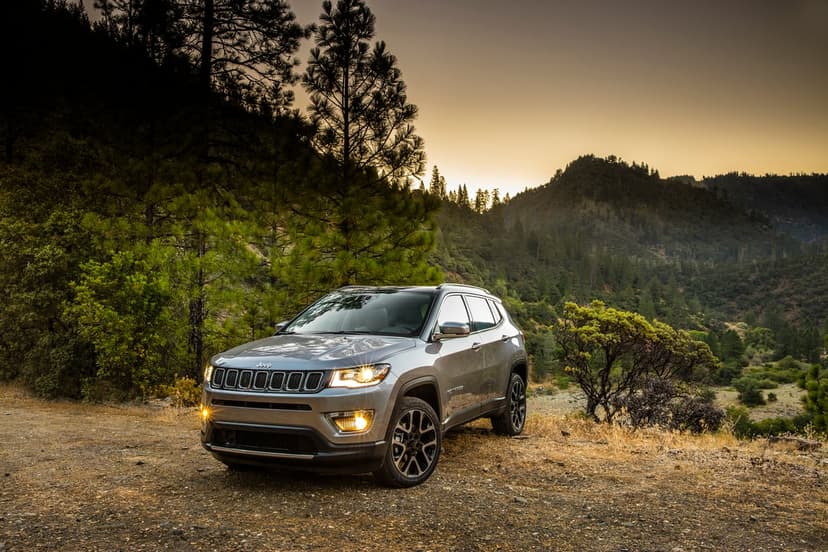 Silver Jeep Compass parked on a dirt path with headlights on against a mountain forest during golden hour.