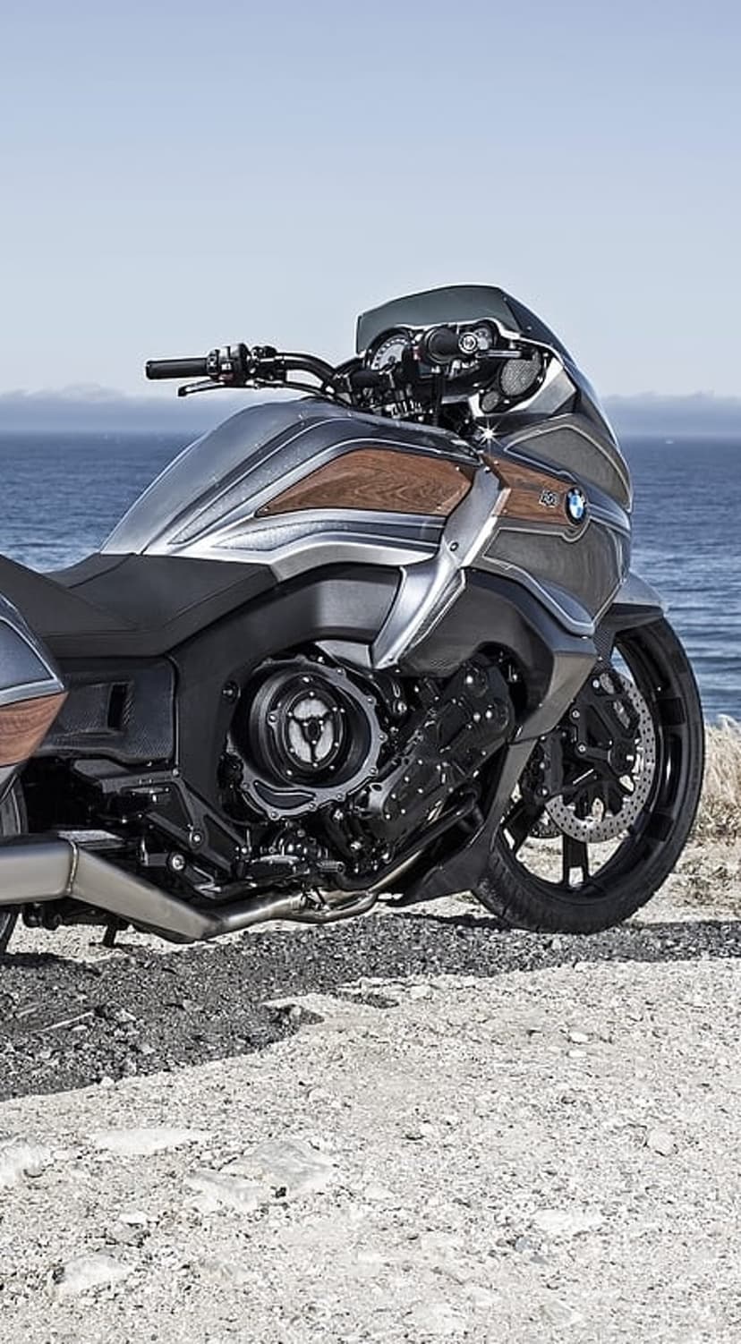BMW Concept 101 motorcycle with wood accents parked on a sandy shoreline under a bright blue coastal sky.
