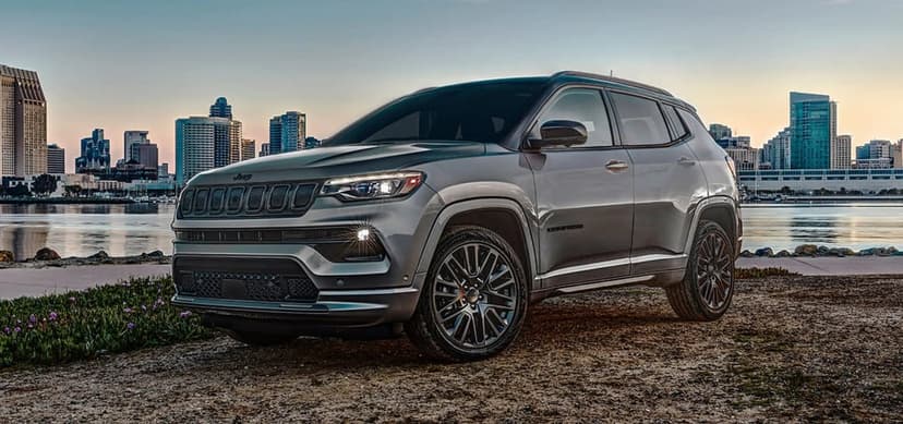 Silver Jeep Compass SUV parked on a gravel shore with a city skyline and calm water under a sunset sky.