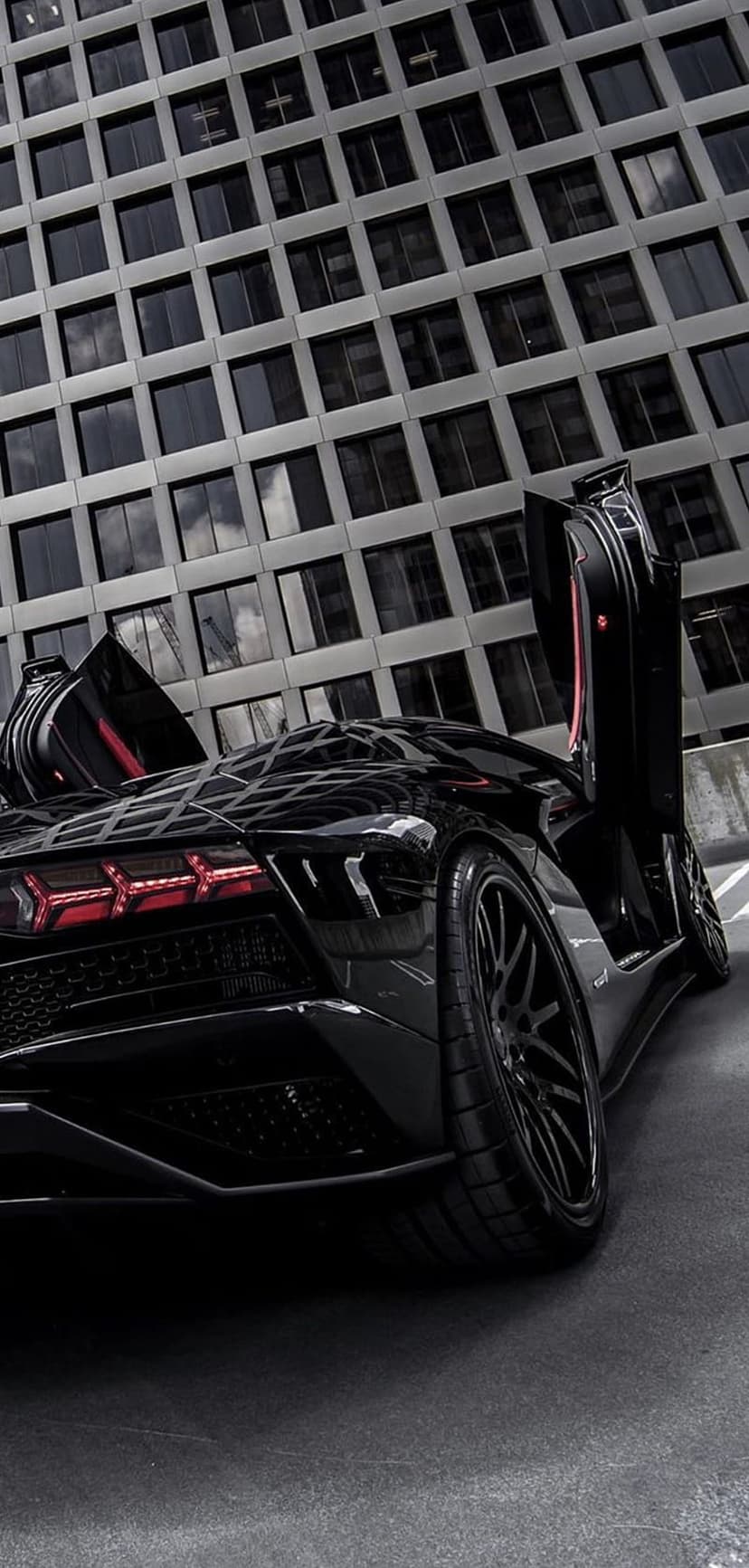 Black Lamborghini Aventador with open scissor doors and red interior accents parked near a grey modern building.