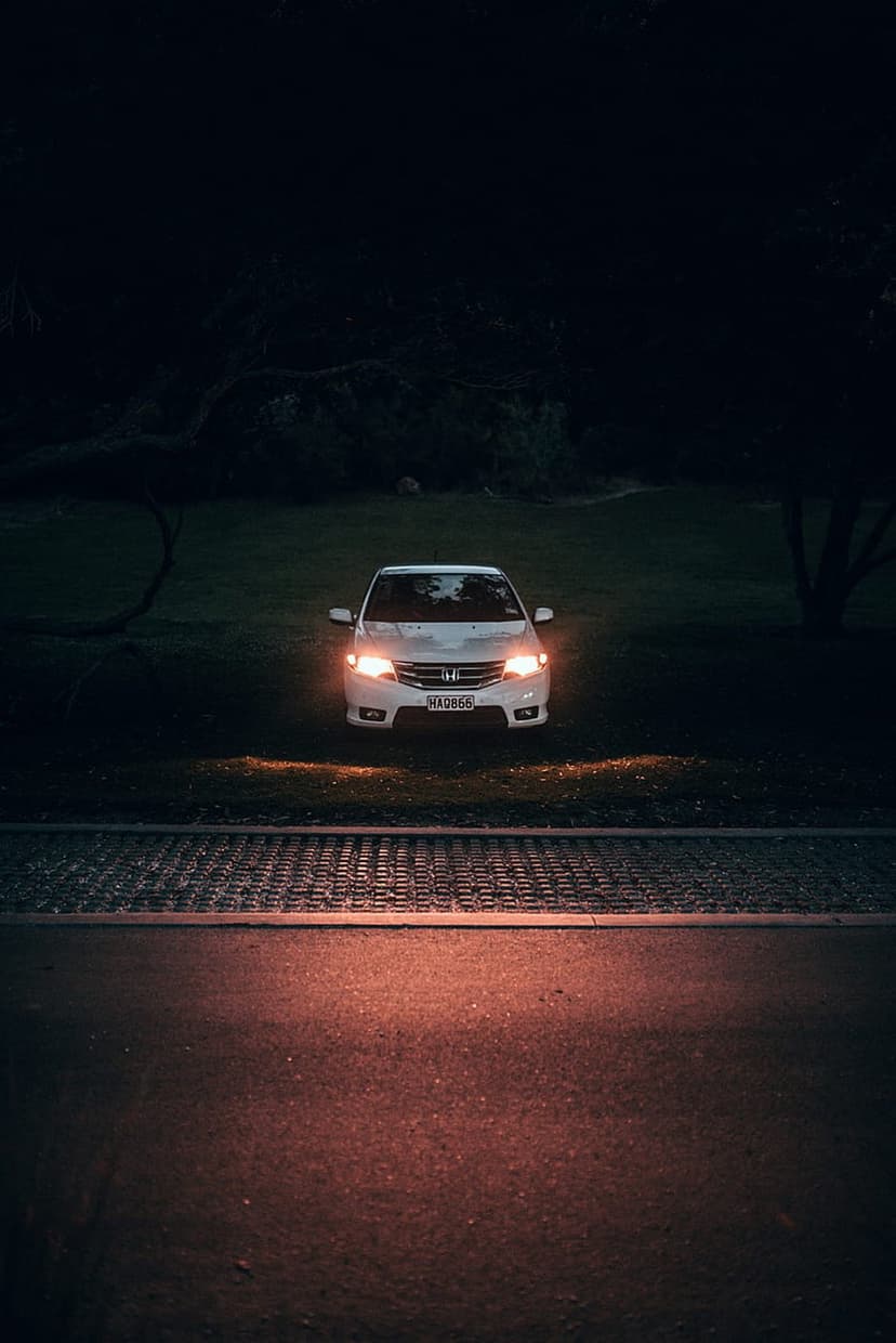 White Honda sedan with glowing headlights parked on grass at night, reflecting warm light on paved ground.