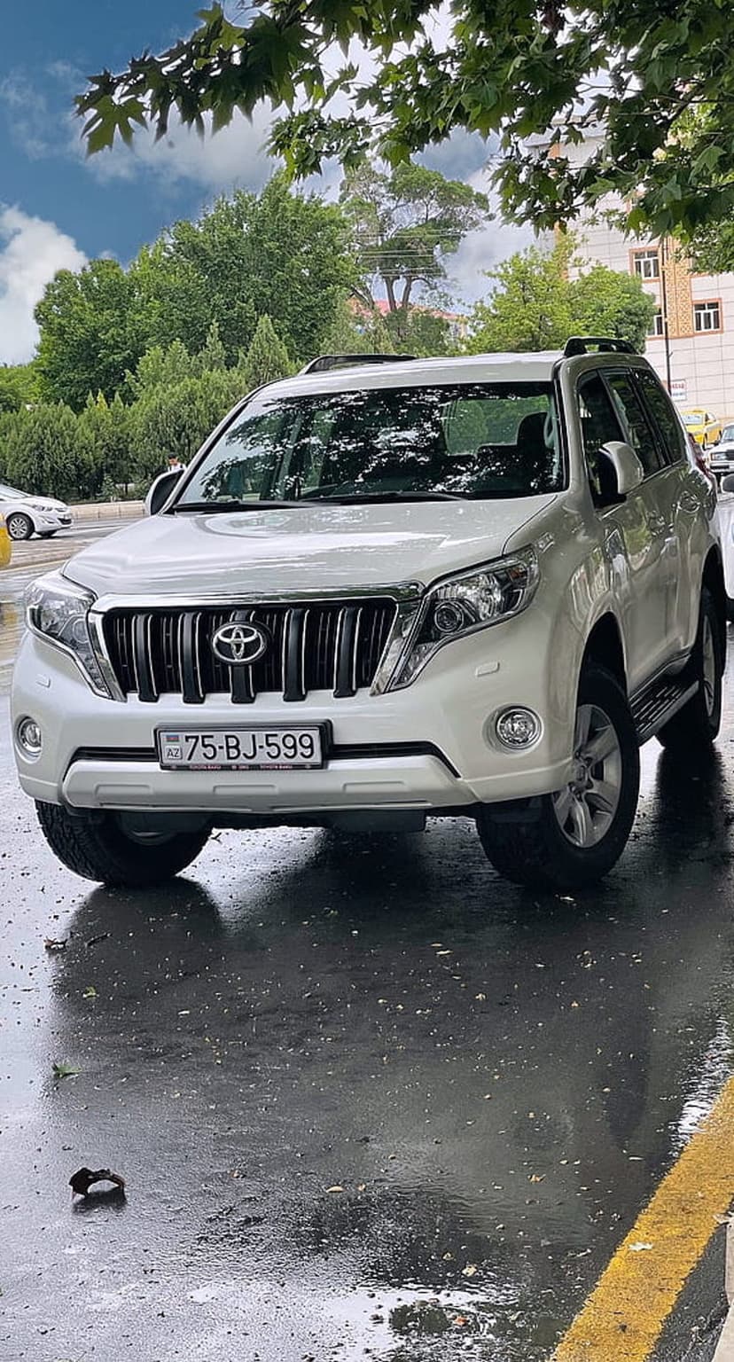 White Toyota Prado SUV parked on wet asphalt with green trees and a cloudy summer sky in the background.