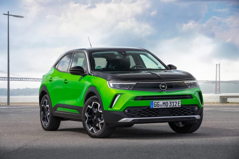 Green and black Opel Mokka-e electric SUV parked on asphalt with a large bridge and cloudy sky in the background.