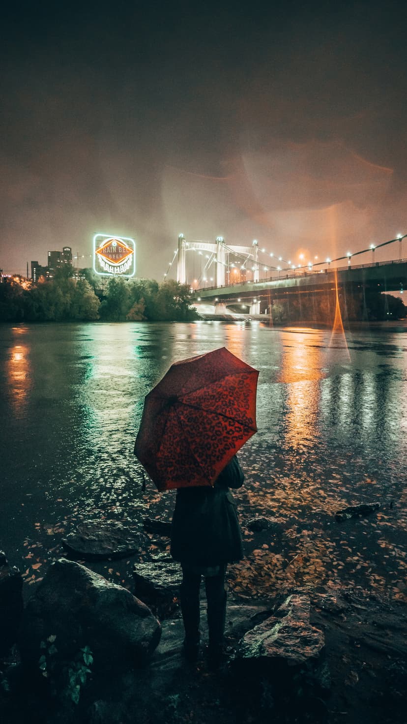 Red umbrella stands on a wet riverside path at night with city lights reflecting in the water under a bridge.