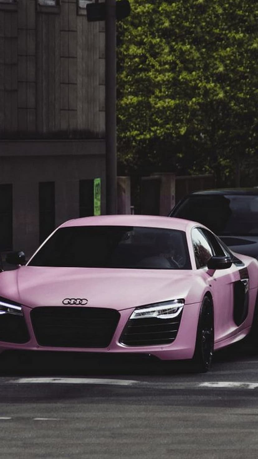Pink Audi R8 supercar with black grille and wheels parked on an urban street with blurred green trees behind it.