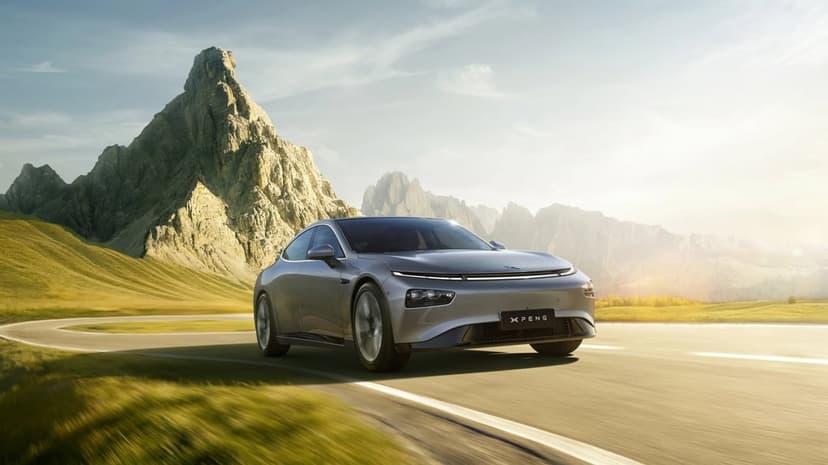 Silver electric sedan driving on a winding mountain road with jagged peaks and golden sunset lighting.