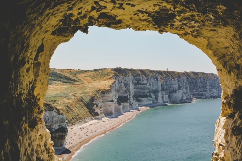Chalk cliffs of Etretat seen from inside a dark sea cave overlooking a pebble beach and turquoise ocean water.