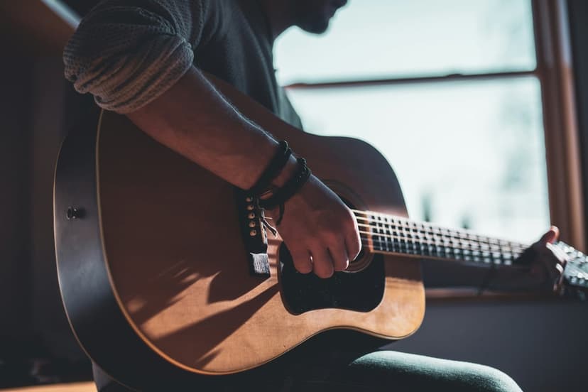 Man playing an acoustic guitar by a sunlit window with dramatic shadows and highlights on the wooden instrument.