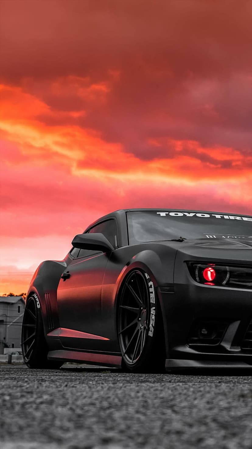 Black widebody Camaro with glowing red halo headlights parked on asphalt during a fiery orange sunset.