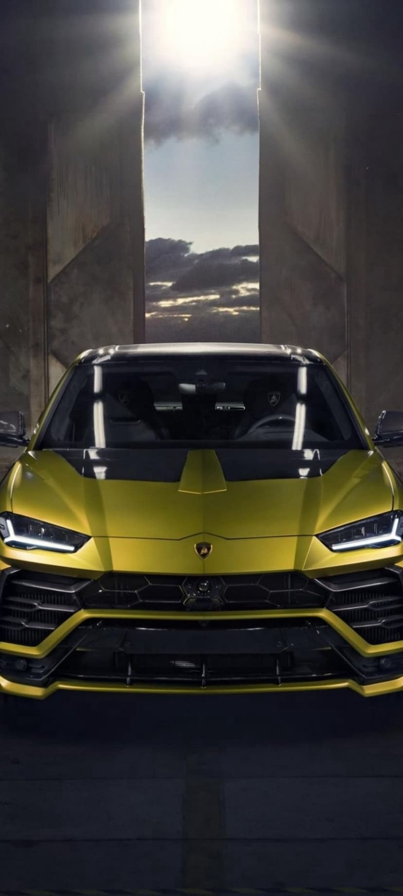 Yellow Lamborghini Urus SUV with sharp headlights emerging from a dark industrial doorway under a cloudy sky.