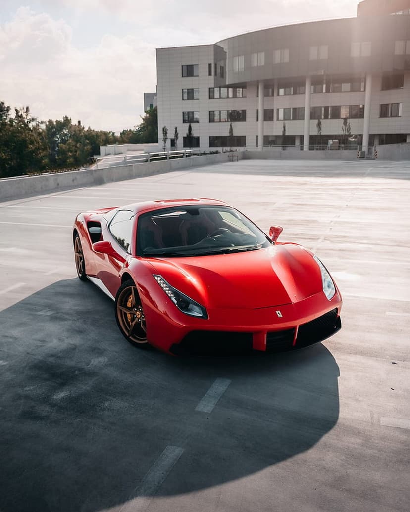 Red Ferrari 488 GTB with gold rims parked on an asphalt rooftop before a curved glass architectural building.