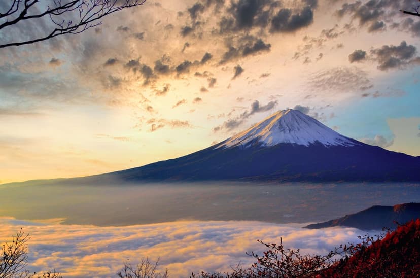 Mount Fuji's snow-covered peak rises above a thick sea of clouds under a glowing golden sunrise in rural Japan.