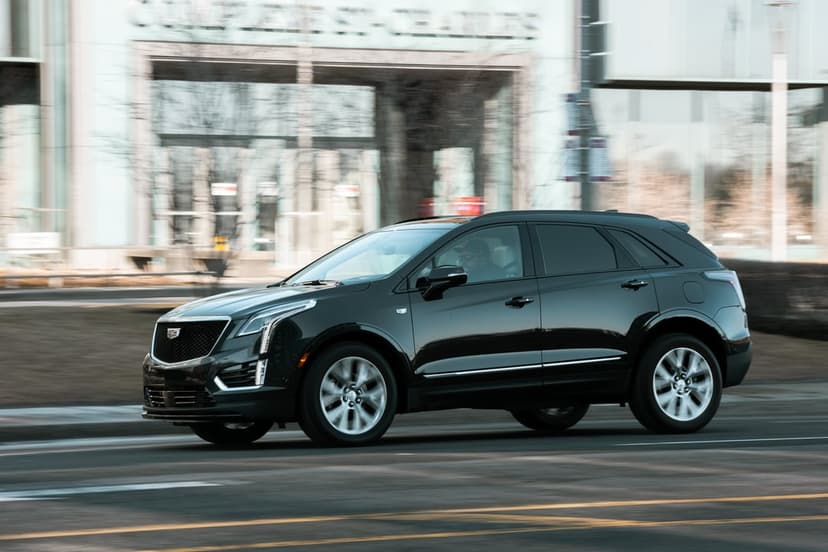Black Cadillac SUV driving on a city street with motion blurred wheels and modern building background.