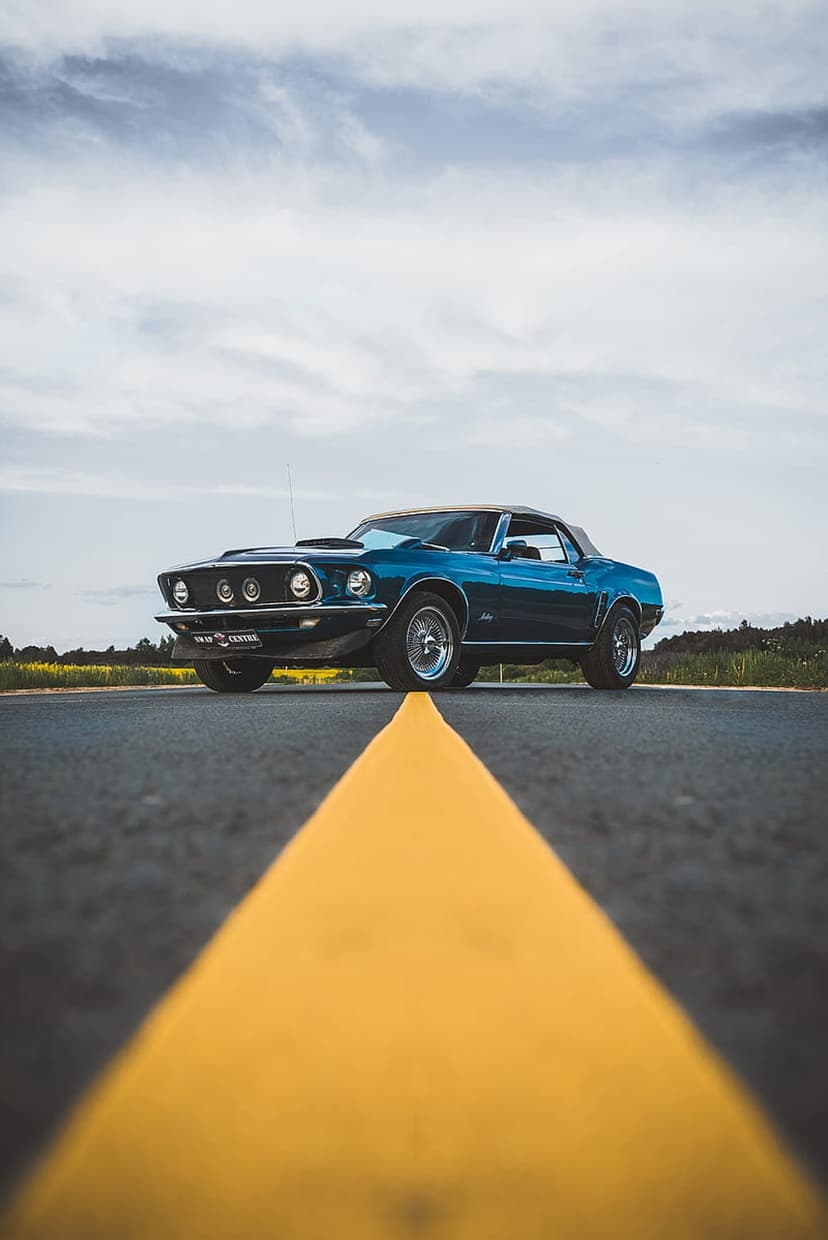 Teal 1969 Ford Mustang convertible on an asphalt road beside yellow flowers under a partly cloudy sky.