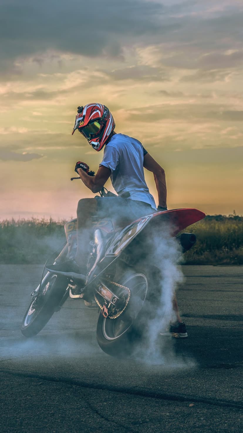 Motorcyclist in a helmet does a wheelie as tire smoke billows across the road under a vibrant sunset sky.
