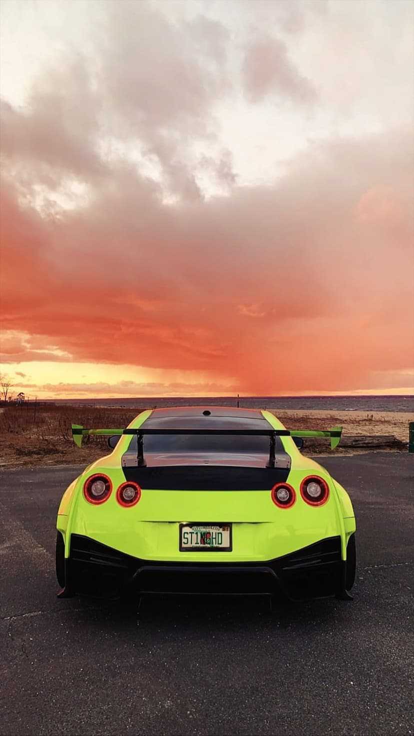 Nissan GT-R in lime green with a large rear wing parked on asphalt against a fiery orange and pink sunset sky.