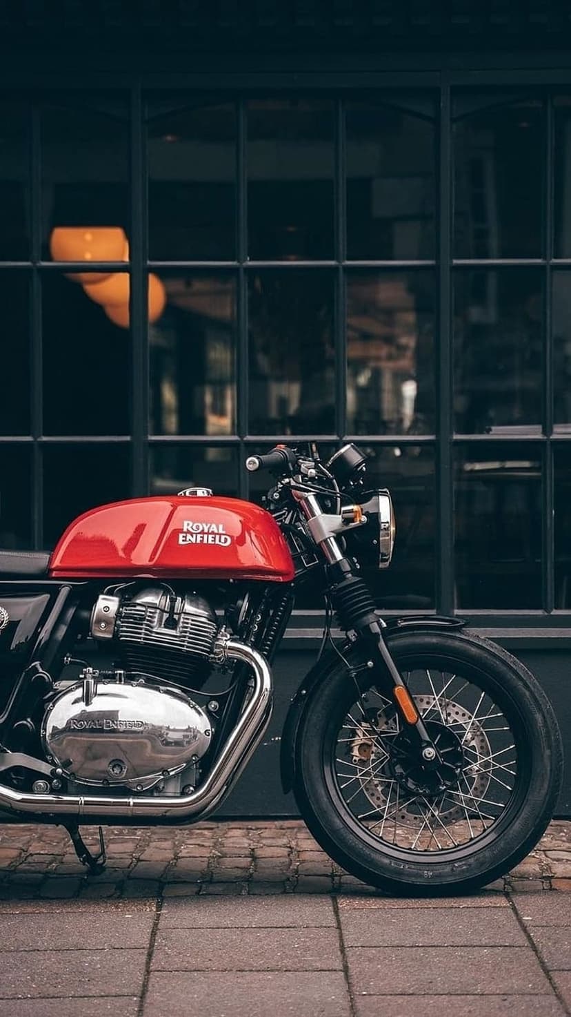 Red Royal Enfield motorcycle with chrome engine parts parked on a brick sidewalk near a glass building facade.