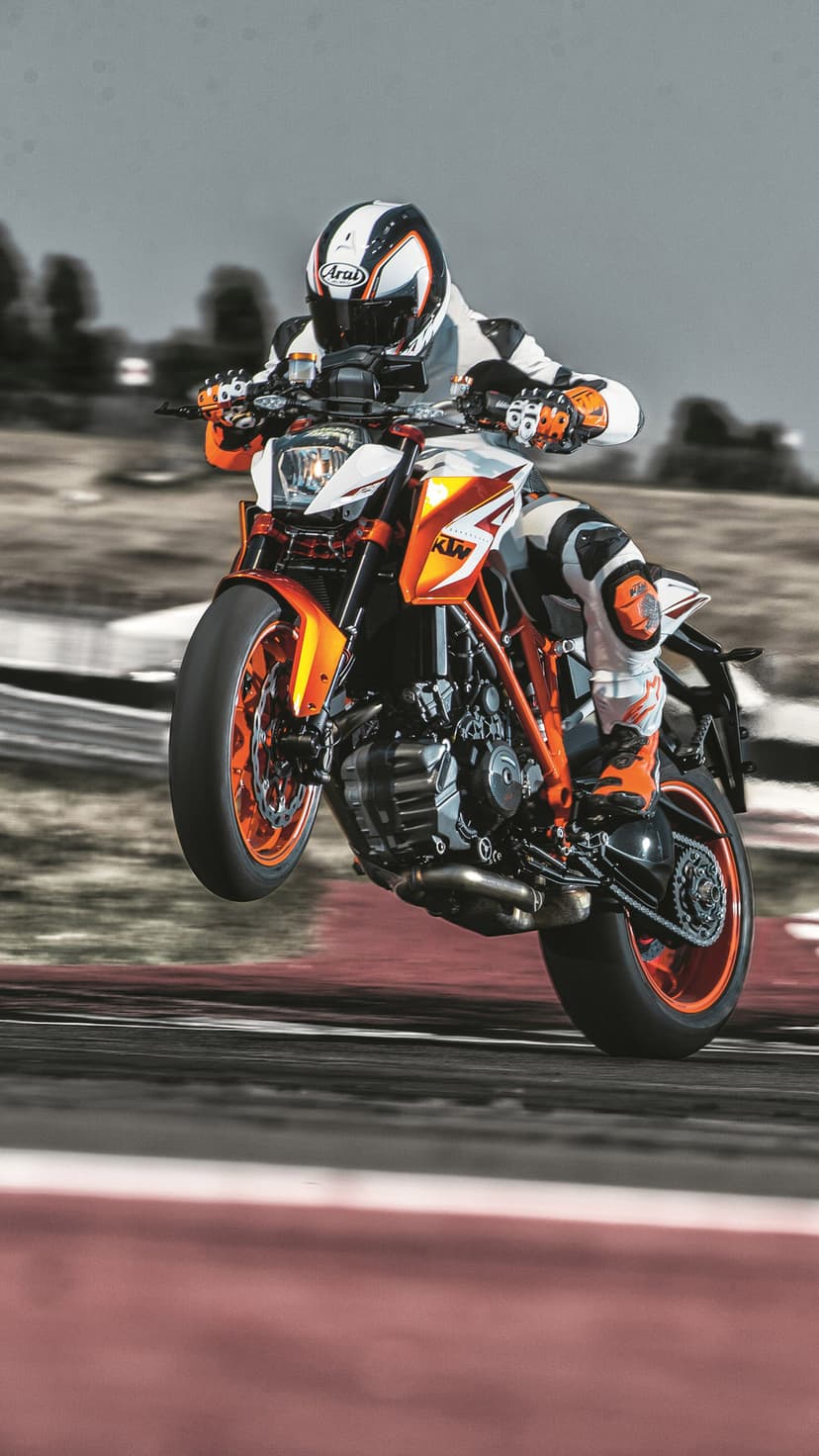 KTM Super Duke R rider in orange leathers performs a front-wheel lift on a blurred asphalt racing circuit.
