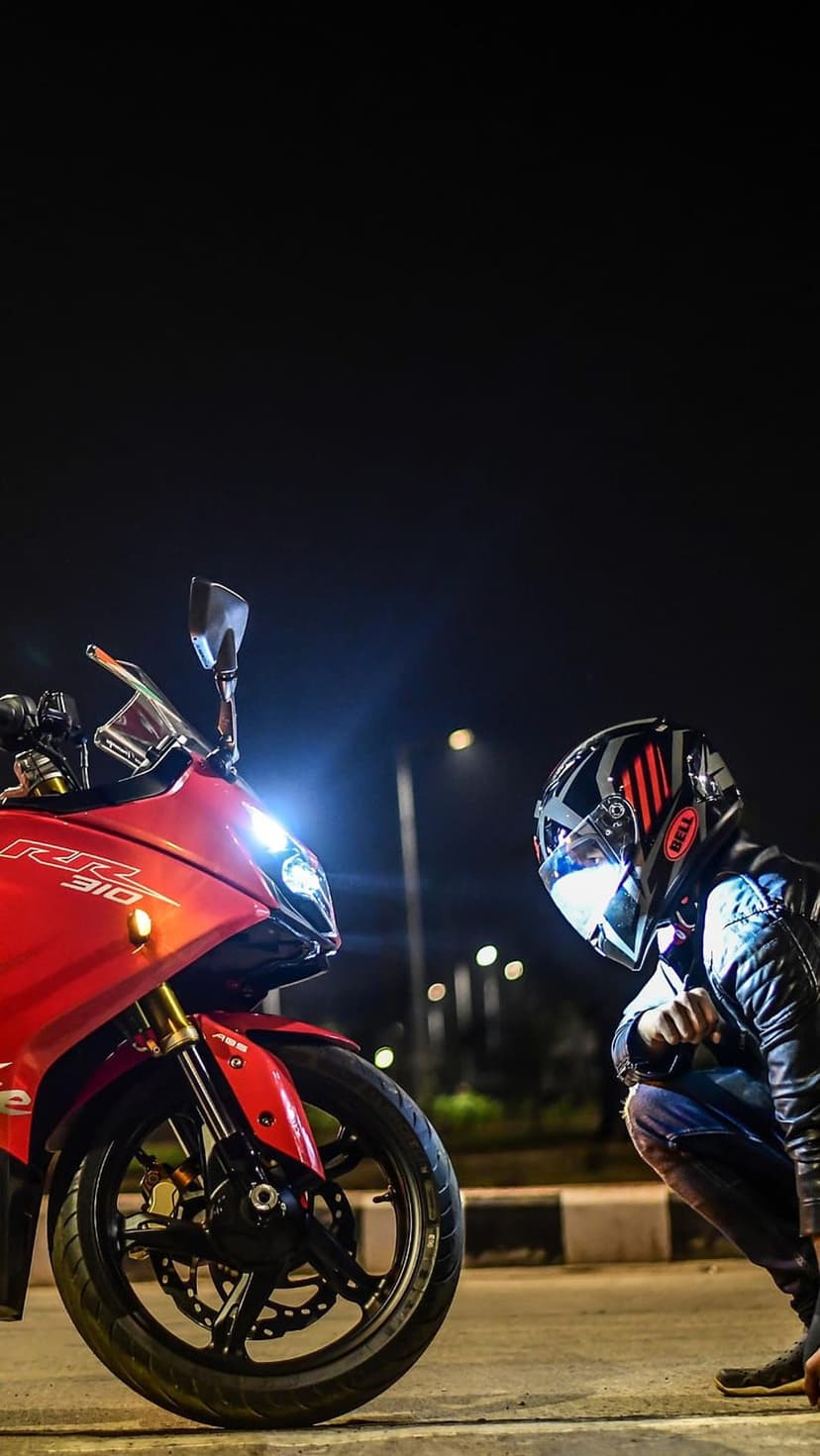Motorcyclist in a leather jacket and helmet kneels next to a red sportbike with glowing headlights on a dark road.