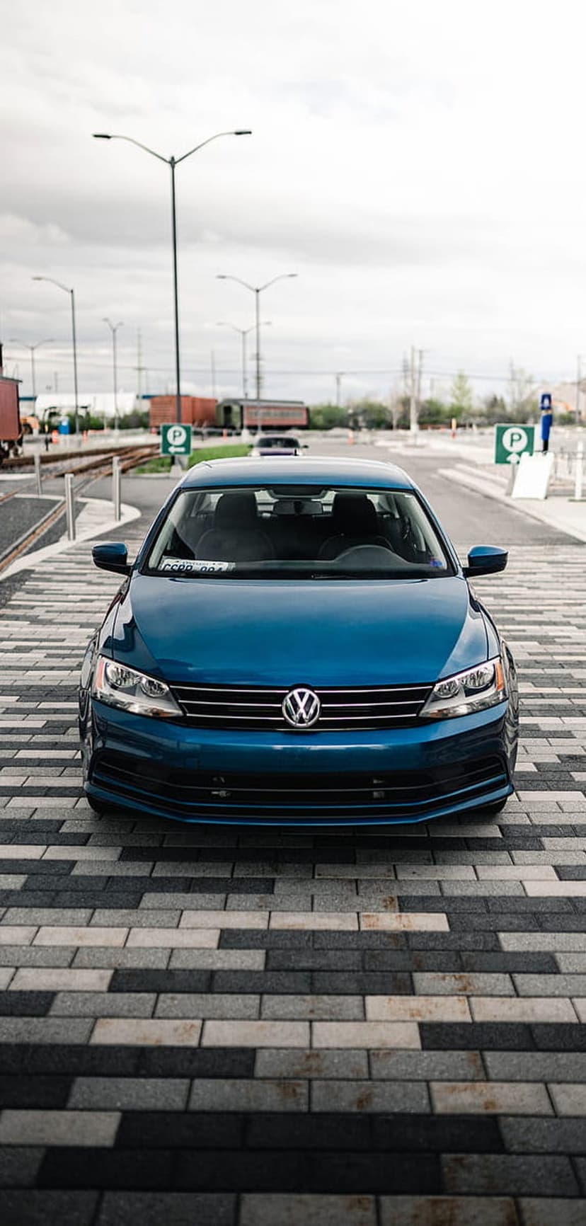 Blue Volkswagen Jetta sedan faces forward on a paved lot beneath industrial streetlights and a cloudy sky.