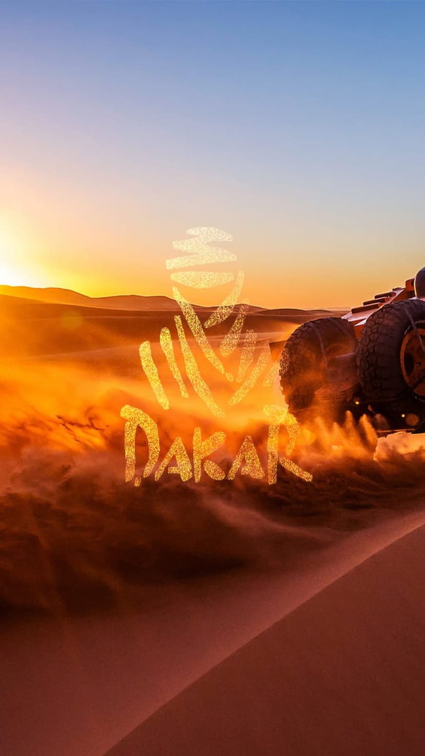Dakar rally car kicks up sand while racing over steep dunes during a vibrant orange sunset with an event logo.