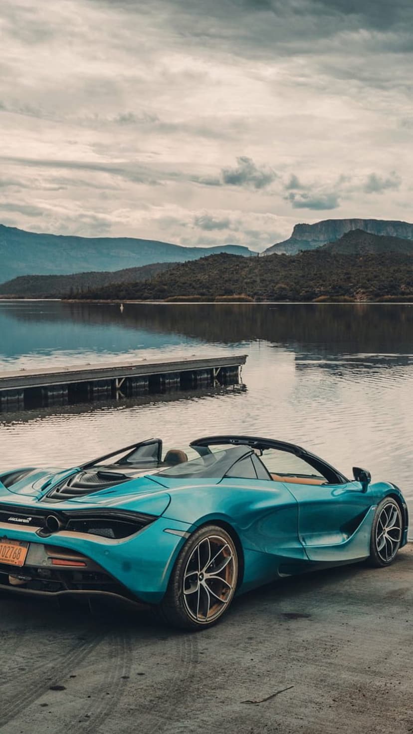 Blue McLaren convertible parked on a lakeside pavement with jagged mountain peaks under a cloudy sky.