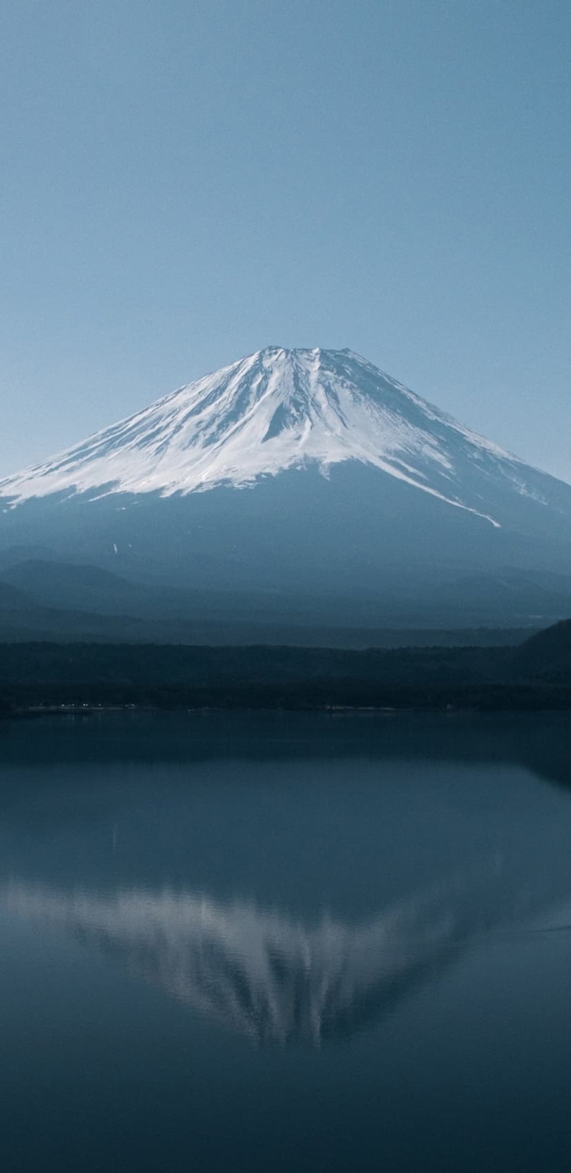 Mount Fuji with a snow-capped peak mirrors in the glassy blue water of Lake Kawaguchi under a clear morning sky.