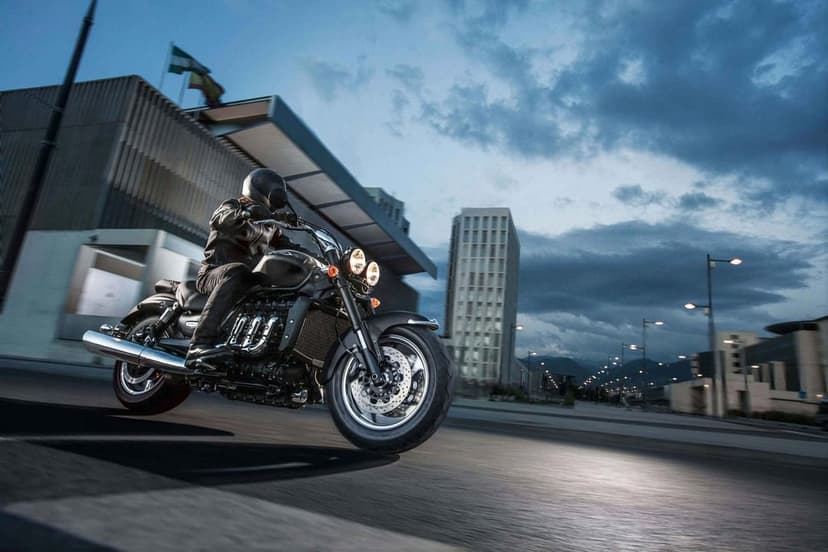 Matte black cruiser motorcycle with dual headlights riding on a city street at night under a cloudy twilight sky.