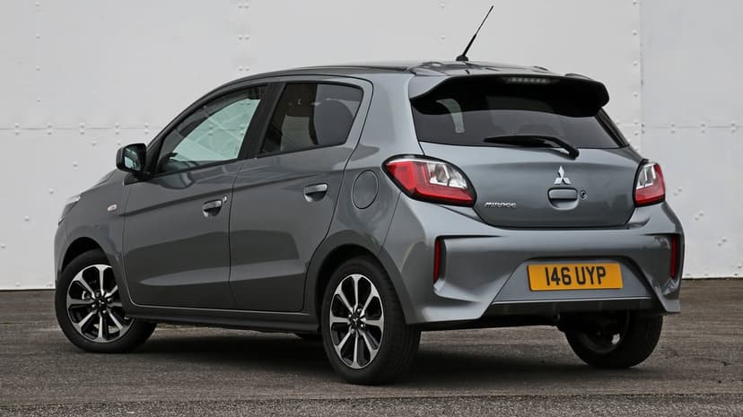 Rear view of a gray Mitsubishi Mirage hatchback with alloy wheels on asphalt against a white riveted wall.