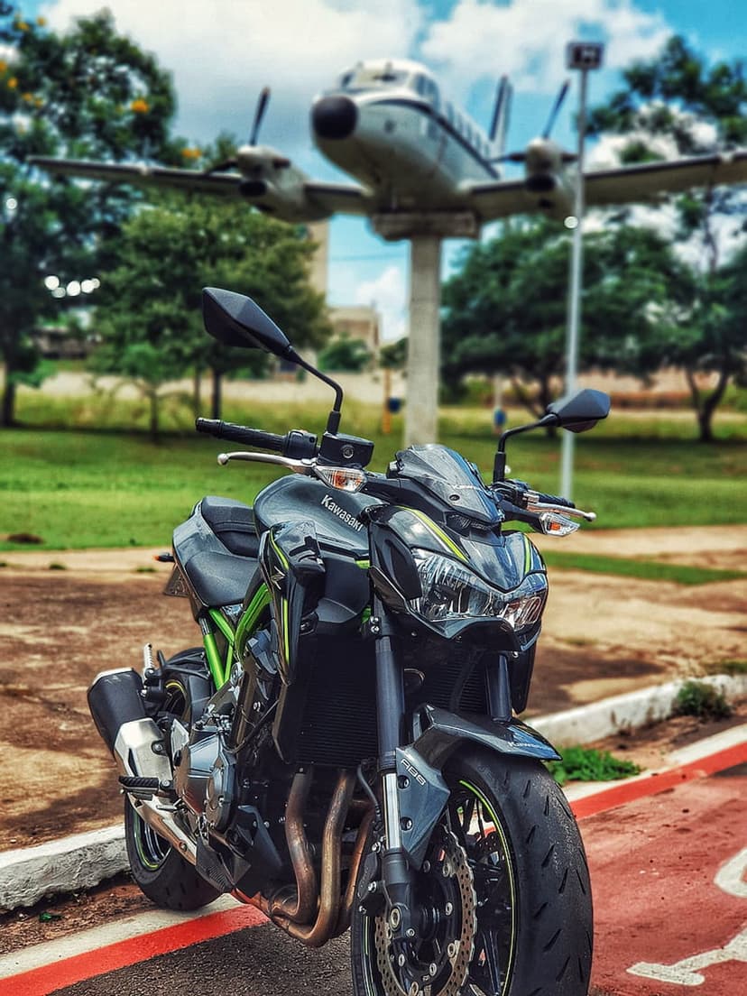 Black Kawasaki motorcycle parked on asphalt with a static aircraft on a pedestal under a clear blue summer sky.