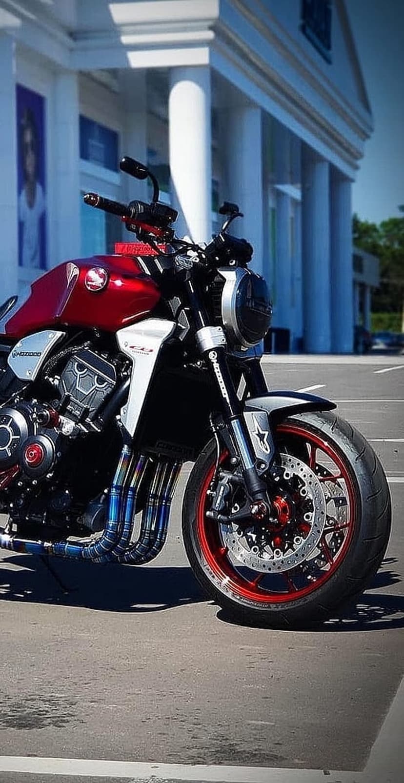 Red custom motorcycle featuring polished chrome exhaust pipes parked outside a modern architectural building.