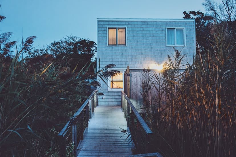 Weathered wooden walkway leads toward a modern blue beach house with glowing windows under a dark twilight sky.