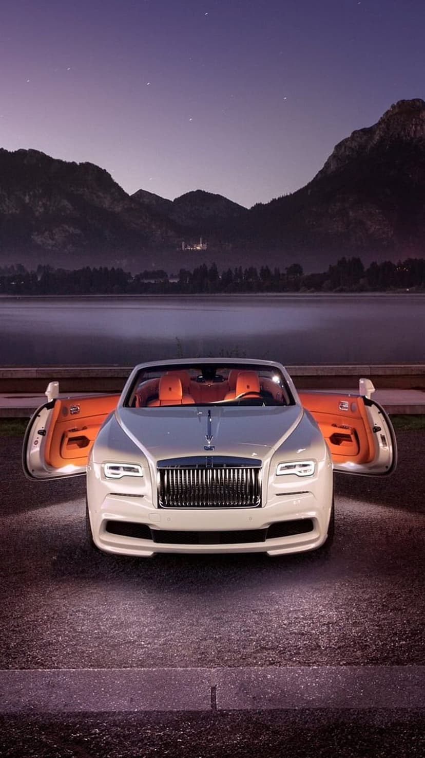 White Rolls-Royce convertible with orange seats parked near a mountain lake under a starry twilight sky.