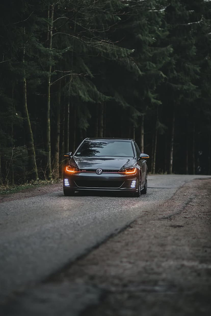 Volkswagen Golf GTI with bright headlights travels along a narrow forest road surrounded by dark pine trees.