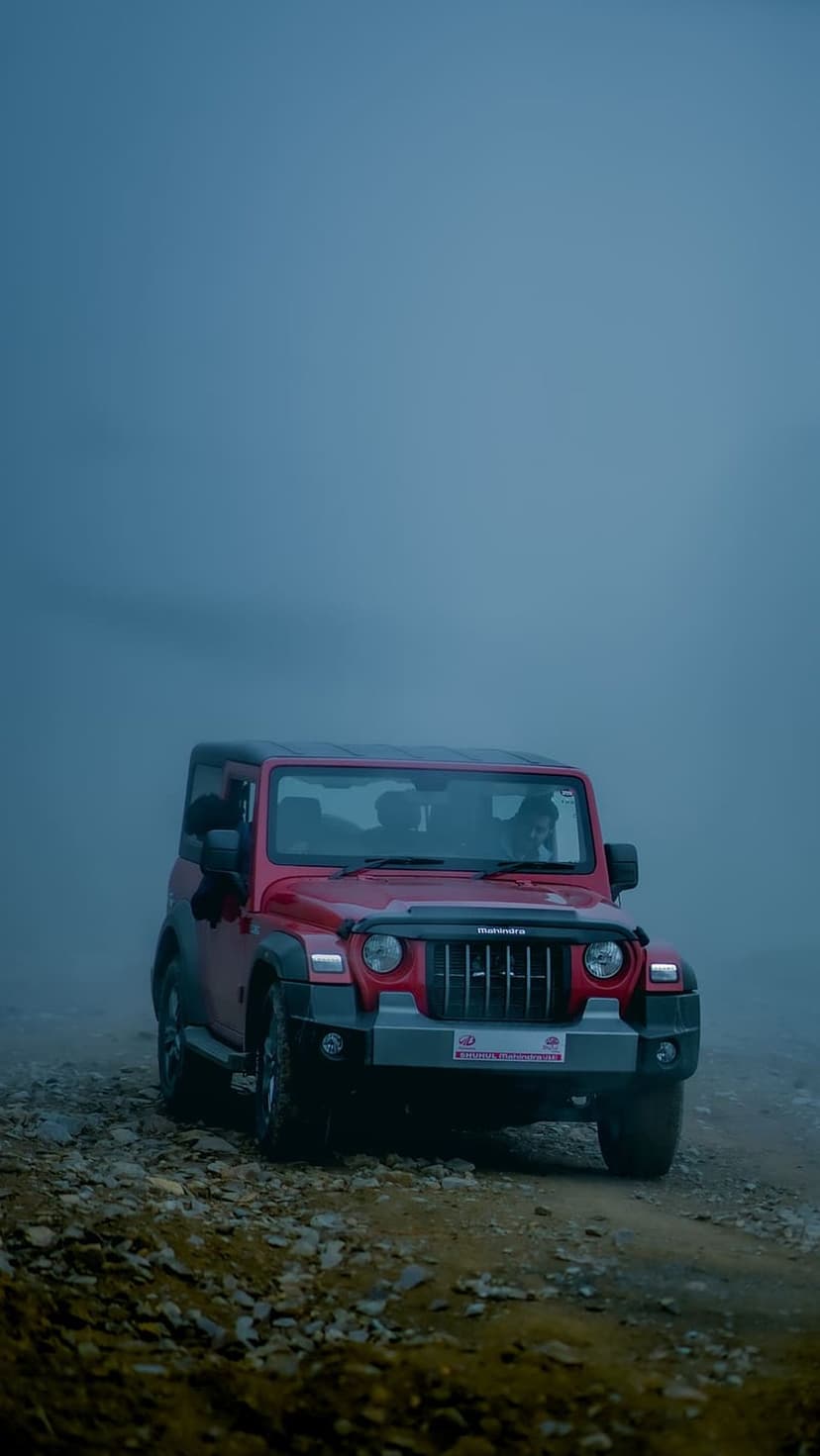 Red Mahindra Thar SUV climbs a rocky mountain path amidst heavy fog and a dim blue sky during an off-road trek.