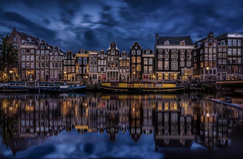 Amsterdam canal night scene shows illuminated historical houses reflecting in dark water under a clear sky.