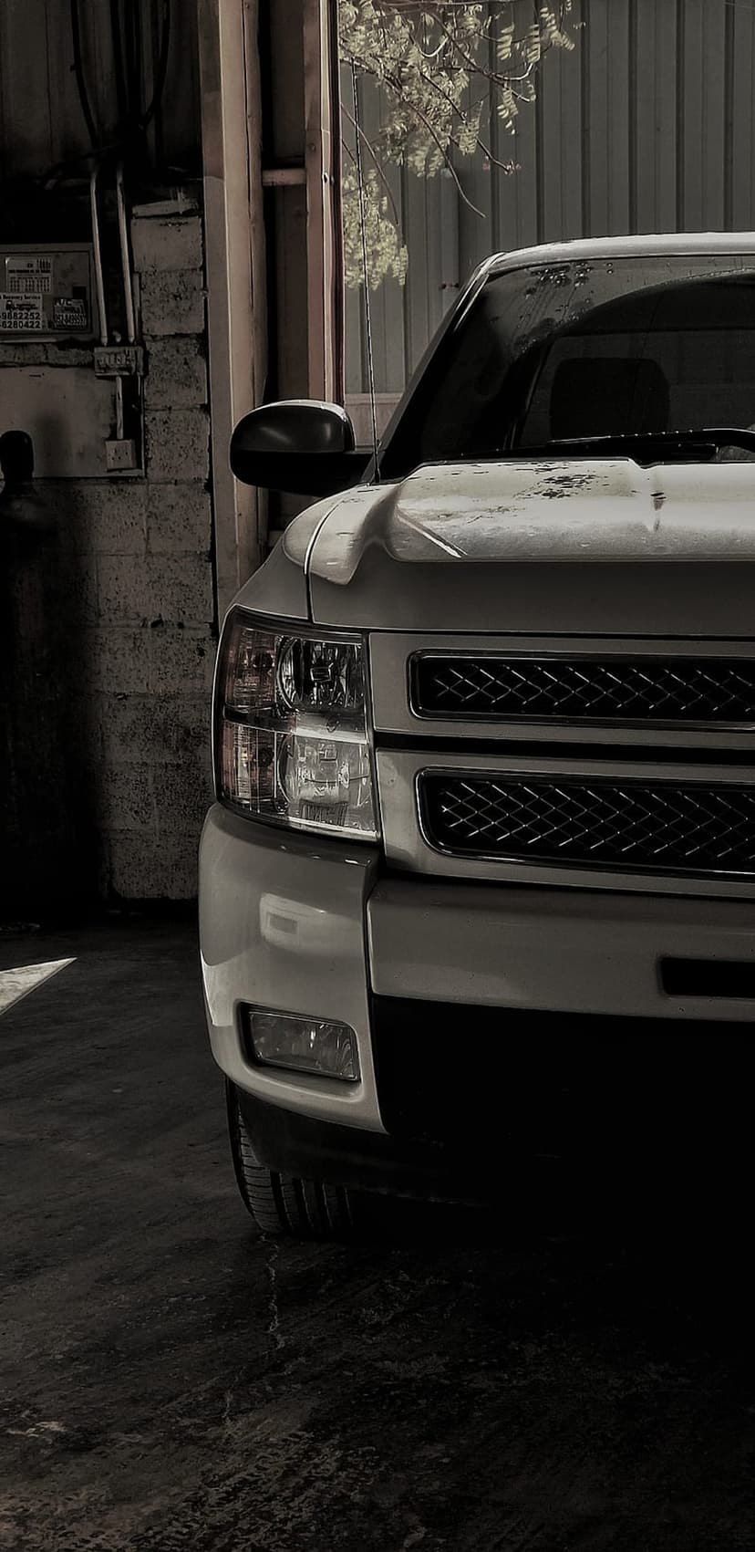 Silver pickup truck front grille and headlight in a dim industrial garage with sunlight on the metallic fender.